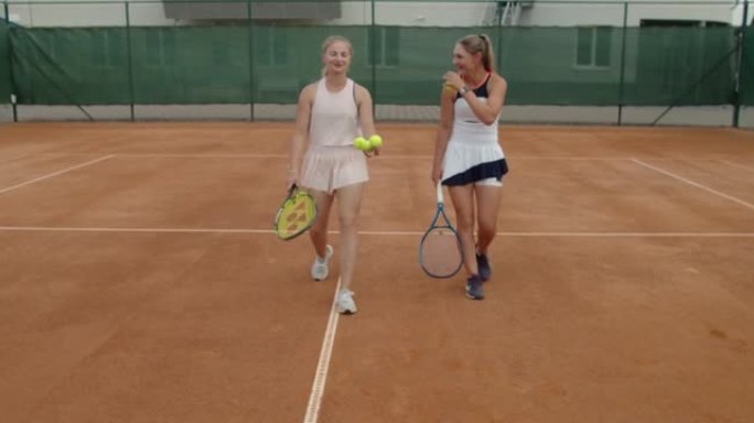 比赛成功后，两个穿着运动服的迷人女孩在网球场上并肩前进。几个朋友球员在训练慢动作后说得很开心。健康健