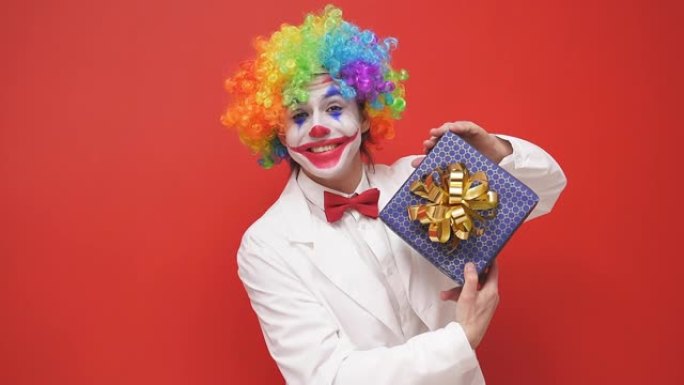 有趣的小丑手里拿着一个礼品盒，在孤立的红色背景下为相机摆姿势