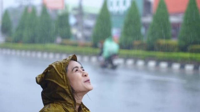 在倾盆大雨的日子里，一个脸上表情积极的亚洲女孩，穿着黄色的雨衣站在路边，在雨中浸湿了，这是亚洲的雨季
