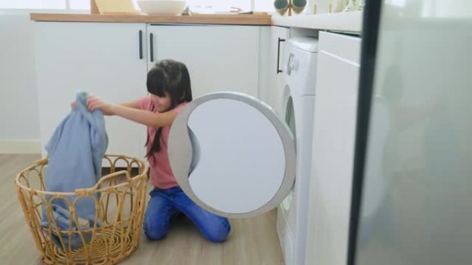 亚洲小孩把脏衣服放进家里的洗衣机里。可爱的女孩孩子坐在地板上，很乐意帮助家人，并在家里的洗衣机里洗衣