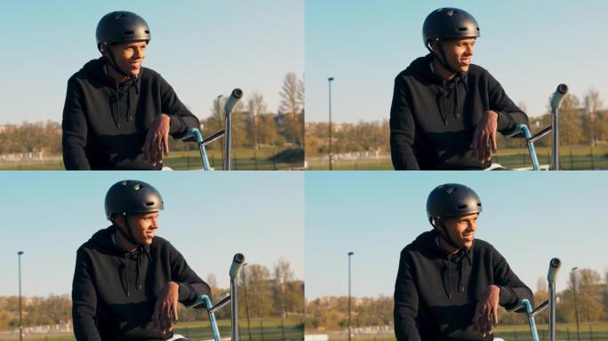 一名年轻的学生面带微笑地坐在滑板场的坡道上，头上戴着头盔，等待着能和他一起骑车和表演特技的同学们