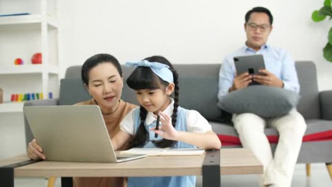 亚洲母亲用笔记本电脑给女儿教作业，而父亲在沙发上放松