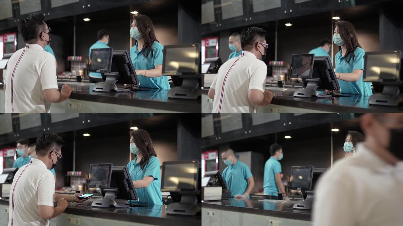 亚洲华人男性顾客戴着口罩，通过手机应用在电影院售票处购买电影票