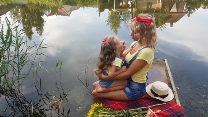 妈妈和小女儿穿着牛仔短裤和背带，头上戴着红色的蝴蝶，坐在河边的钓鱼平台上，拥抱亲吻