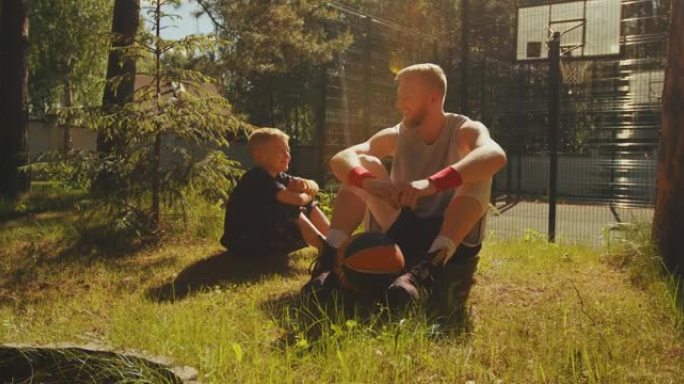 运动的年轻父子篮球运动员坐在草坪上与球交流。快乐育儿理念