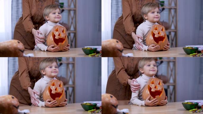 这个男孩坐在桌子旁，看着前方，拥抱着一个大的橙色南瓜。孩子开始对某事感到高兴，他的母亲站在他身后抚摸