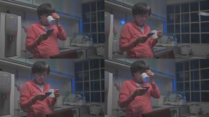 亚洲华裔高级妇女在晚上用智能手机查看短信，在家庭厨房喝水