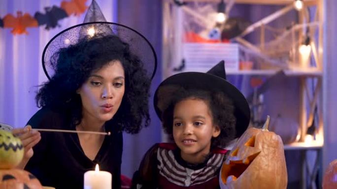小女孩和女人，戴着巫婆帽的非洲裔美国人，用雕刻的笑脸点燃南瓜蜡烛。妈妈和女儿坐在万圣节之夜装饰的房间