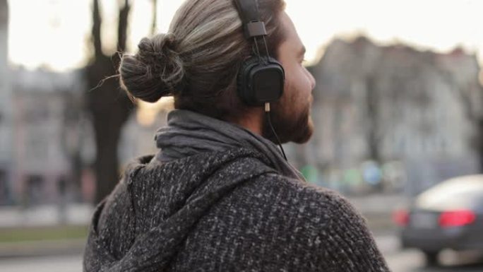 一个大胡子的男人正戴着耳机听音乐，在城市里走来走去。他的头发被绑起来了。从侧面拍摄肖像。他正在四处张