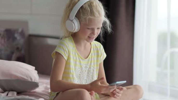 戴着耳机的金发小女孩，手里拿着手机。孩子在听音乐。在家休息，放松，安静的时间