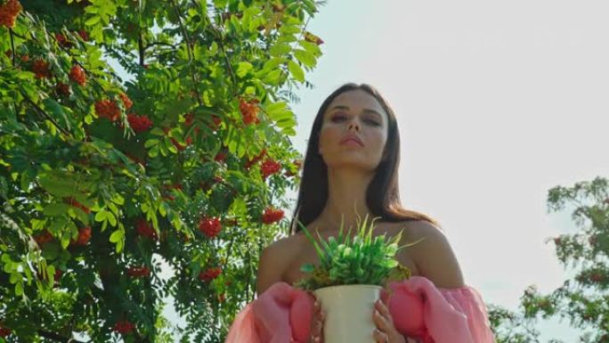 迷人的年轻女子在公园、森林或花园中拿着花盆靠在树上。裸露肩膀的女孩站在一棵花椰树下，树丛着橙色的水果