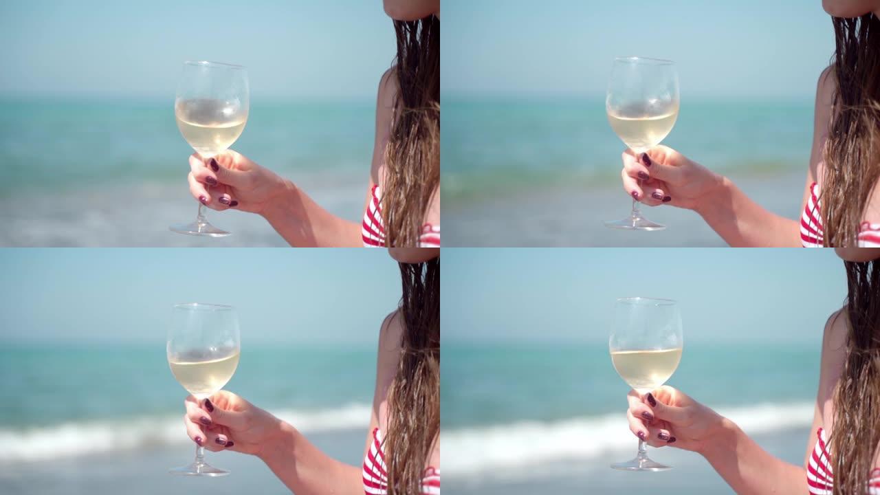 女人的手的特写镜头，一杯新鲜的白葡萄酒在令人惊叹的海景背景和即将来临的海浪中，漂亮的女性在慢动作中挥