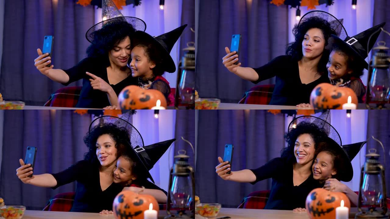 小女孩和年轻女子戴着巫婆帽子的非洲裔美国人使用智能手机通过视频通话进行交流。妈妈和女儿坐在万圣节之夜