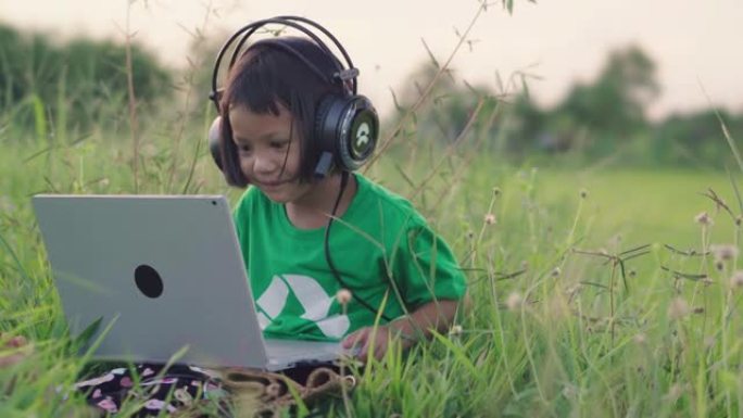 小女孩戴着耳机，在绿草丛中使用笔记本电脑进行社交媒体。农村场景。在夏天放松时间