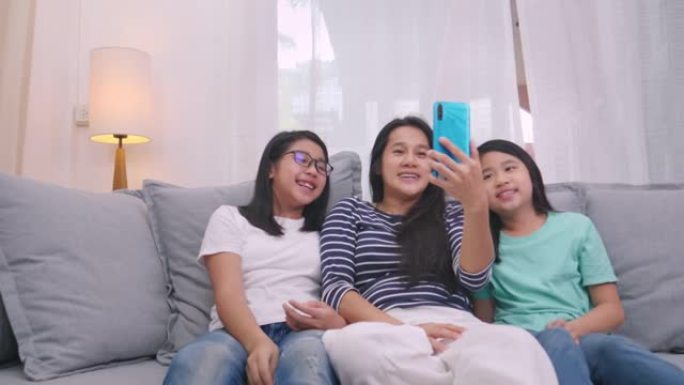 幸福的家庭坐在客厅的沙发上与某人一起通过手机交谈，微笑的母亲通过智能手机向可爱的小孩视频通话与家人交