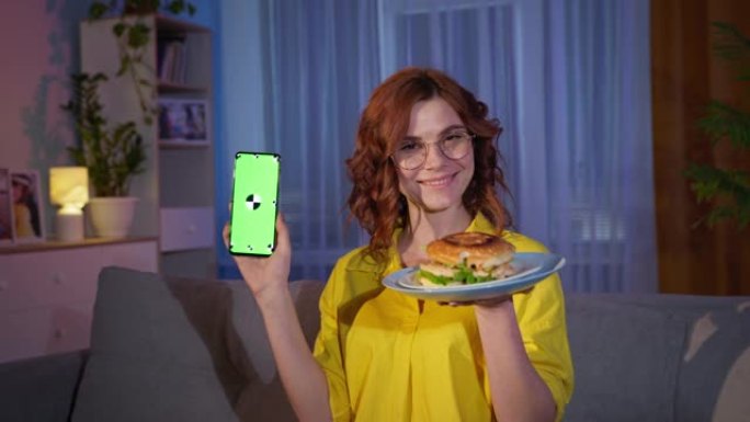 在线订购快餐，戴着汉堡和手机的快乐女人，手里拿着绿色色度键，在家里吃晚饭时对着相机微笑和手表
