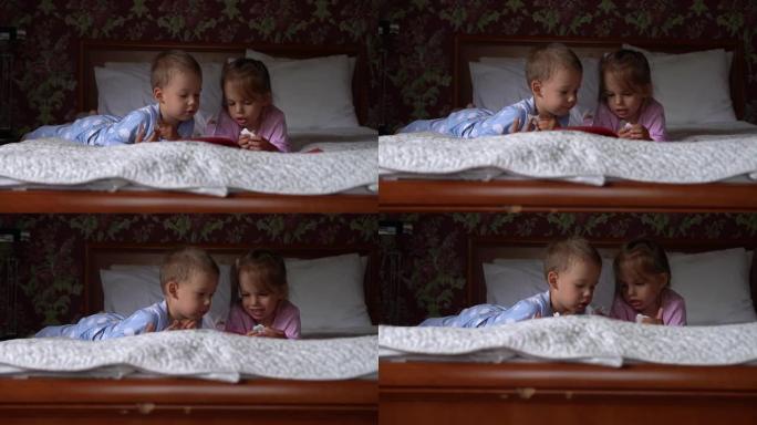 两个可爱的微笑的preshool，穿着睡衣的蹒跚学步的孩子在床上读童话书。兄弟姐妹小双胞胎读者玩得开