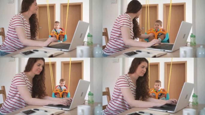 快乐的母亲在家庭办公室里用笔记本电脑工作时摇摆着男婴