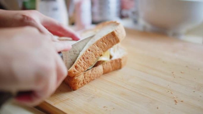 近距离的人在厨房的木板上用锋利的刀将肉，奶酪和蔬菜的三明治面包切成两半，在家中制作快餐