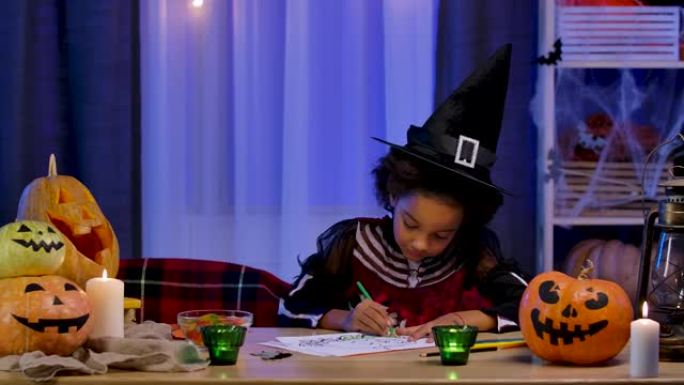 穿着节日服装和巫婆帽子的非洲裔美国小女孩用铅笔画南瓜。孩子坐在万圣节之夜装饰的房间的桌子旁。慢动作