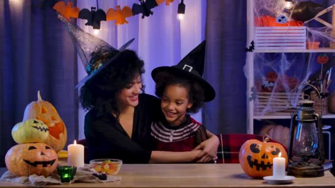 妈妈和女儿穿着节日服装和巫婆帽子的非洲裔美国人玩得开心，快乐地拥抱。幸福的家庭坐在万圣节之夜装饰的房