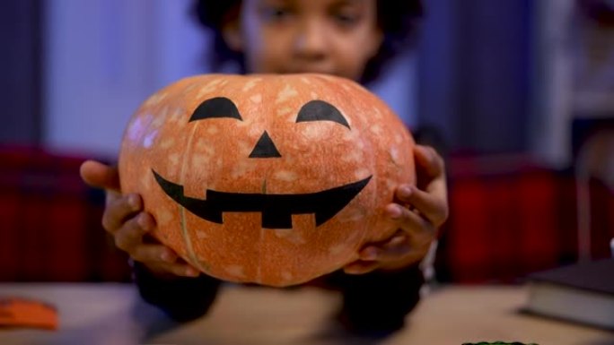 穿着节日服装的非洲裔美国小女孩拿着一个橙色的南瓜，脸上挂着微笑的脸。孩子坐在万圣节之夜装饰的房间的桌