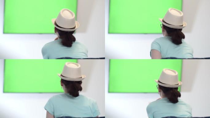 戴帽子的女孩坐在Geen Sreen电视的字体中，切换频道并吃芯片，专心观看电视上的信息，绿色色度键
