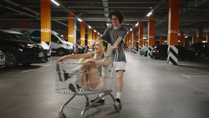 两个时髦的人在地下停车场鬼混，家伙骑着他的女朋友坐在购物车里，