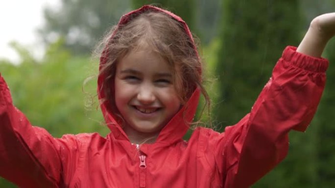 快乐的小女孩在雨中微笑。孩子热爱自然，幸福自由。玩水滴雨的孩子。儿童在下雨的夏日在户外玩耍。穿着红色
