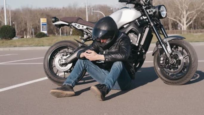 年轻迷人的摩托车手，戴着黑色头盔和黑色皮夹克，在街上使用智能手机