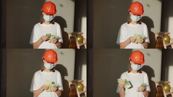 年轻的微笑女工戴着橙色安全帽和医用口罩数钱，站在公寓的建筑工地。建筑师持有现金用于房屋处理