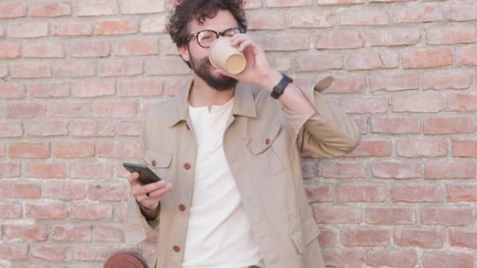 一个不经意的男人靠在砖墙上，举着咖啡喝着，拿着电话，戴着眼镜