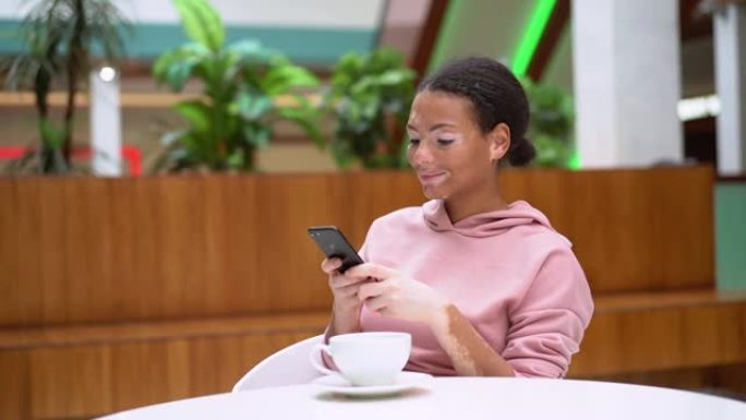 患有白癜风色素沉着皮肤问题的黑人非洲裔美国女性室内穿着粉色连帽衫使用智能手机