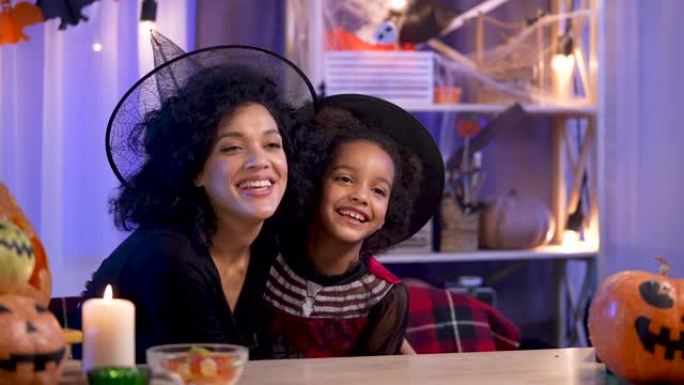 妈妈和女儿穿着节日服装和巫婆帽子的非洲裔美国人玩得开心，快乐地拥抱。幸福的家庭坐在万圣节之夜装饰的房
