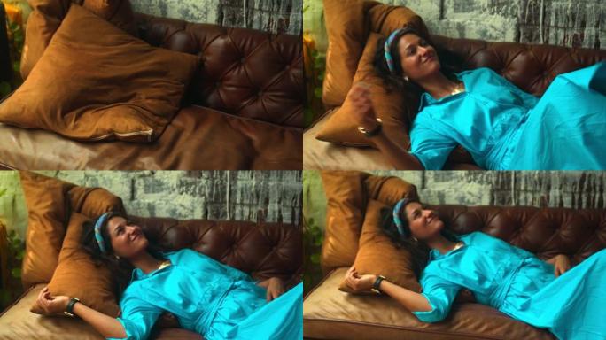 穿着蓝色连衣裙的美丽欧洲黑发女孩躺在沙发上，用金戒指抚摸她的手