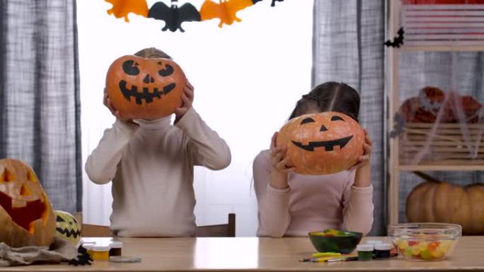 两个孩子，一个男孩和一个女孩，在他们面前举起南瓜，脸上涂着可怕的面孔。他们把南瓜放在桌子上做鬼脸。孩