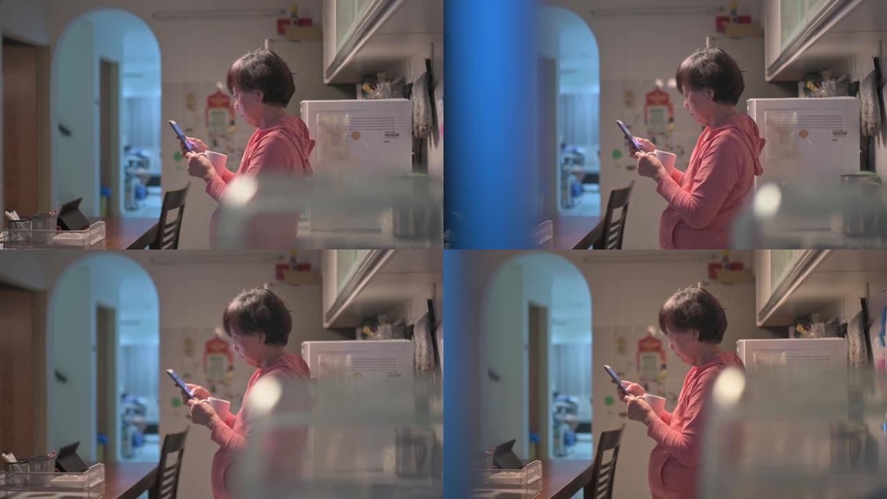 亚洲华裔高级妇女在晚上用智能手机查看短信，在家庭厨房喝水