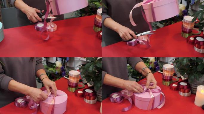 关闭卖家手在礼品店的圆形礼物盒上绑上粉色胶带。女性手在商店里用丝带包装圆形粉色盒子。