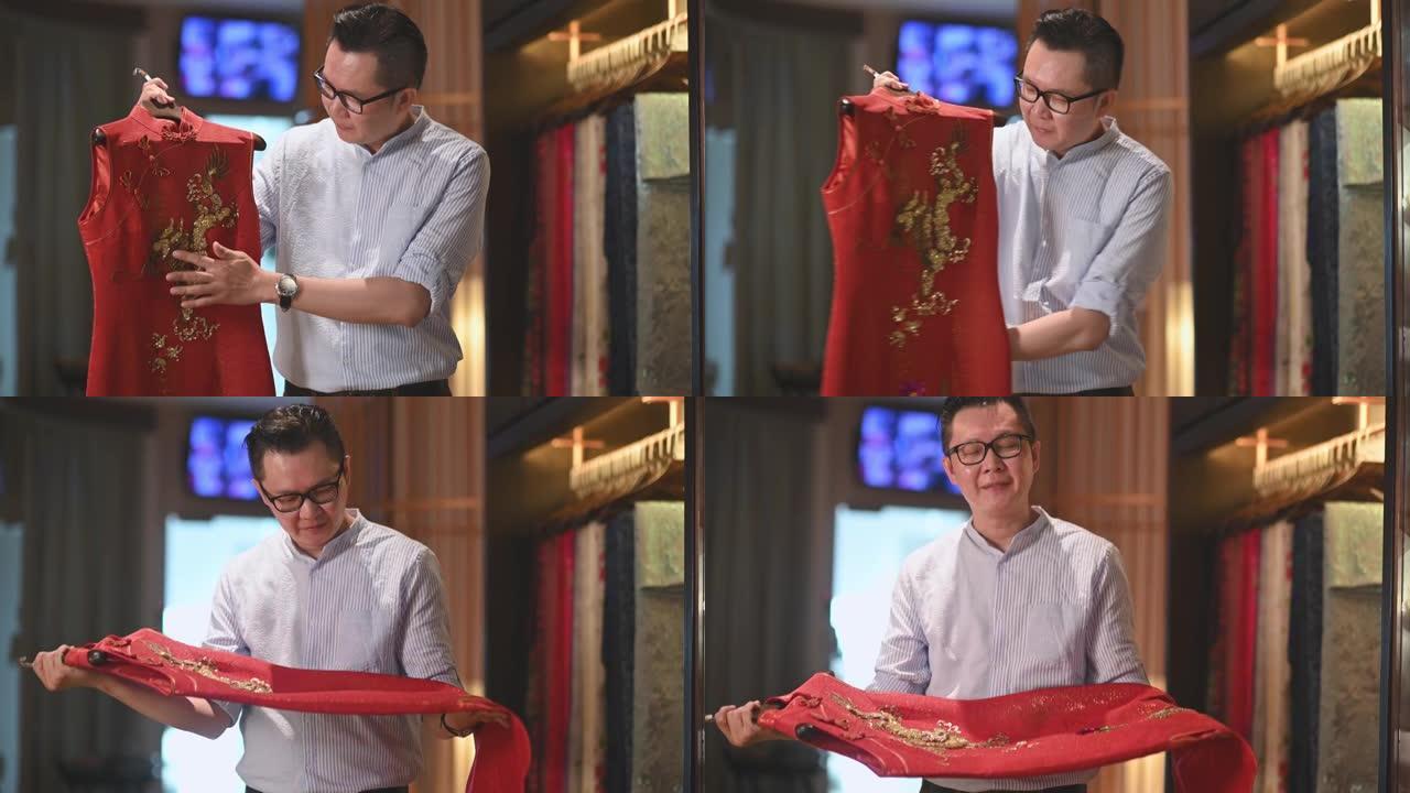 自信的男性中国传统服装时装设计师拿着和看着传统服装旗袍在他的设计工作室精品店微笑着