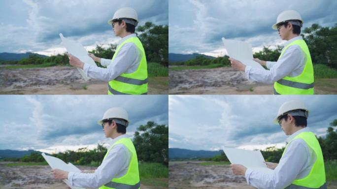 亚洲男工程师穿着安全背心和头盔看建筑工地的建筑蓝图，安全工程建筑专家职业，房地产建筑规划师