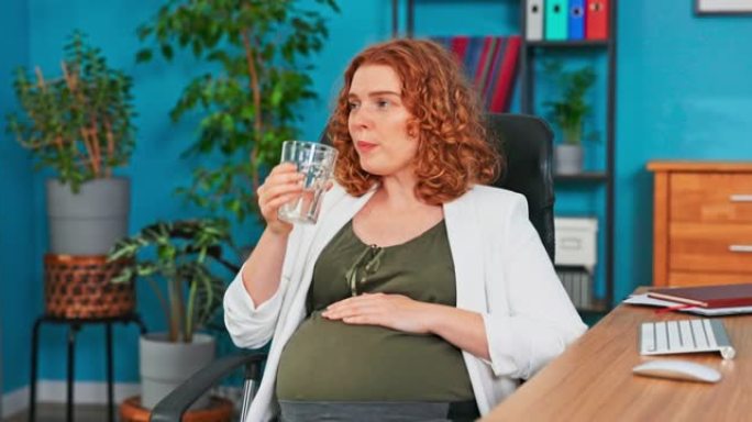 准妈妈在电脑显示器前的办公桌上工作，在转椅上休息，在怀孕的肚子上抚摸自己，喝水，补水，照顾健康后，感