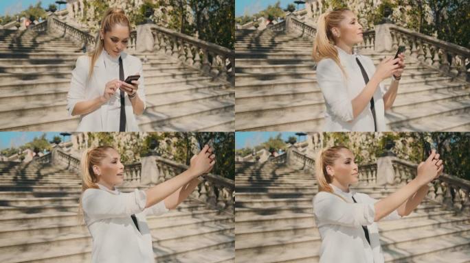 经典套房中年轻美丽优雅的女人自信地在智能手机上自拍站在老城区公园的楼梯上的建筑