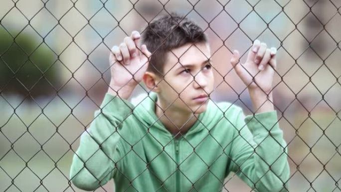 难民悲伤的男孩独自一人低头在篱笆附近，垂下眼睛，拍摄电影