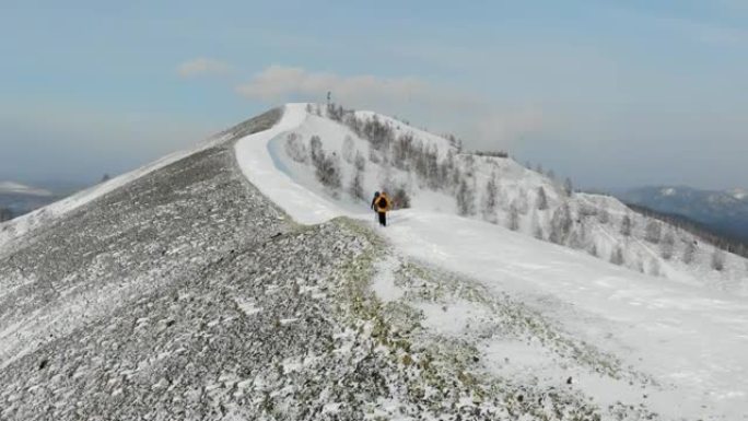 男人和女人正沿着白雪覆盖的山顶上的小路行走。穿着黄色夹克的男人正用棍子走路。北欧行走。你可以看到远处