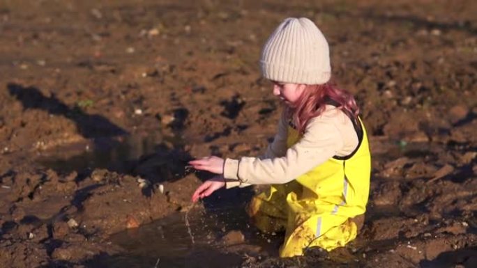 可爱的孩子坐在泥泞的水坑里玩耍。春天在村子里的小女孩。快乐的童年女儿。森林里积雪融化的湿地面。黄色连