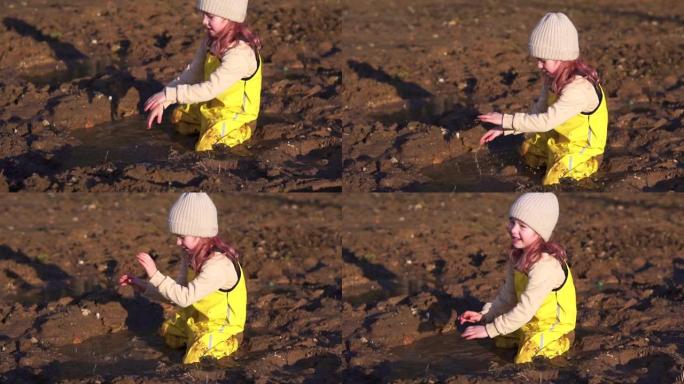 可爱的孩子坐在泥泞的水坑里玩耍。春天在村子里的小女孩。快乐的童年女儿。森林里积雪融化的湿地面。黄色连