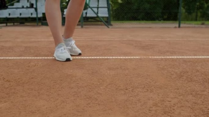 网球运动员弹跳球的低角度裁剪视图，将球从红土球场慢动作中抬起。近距离女性双手拿起球进行户外运动练习。