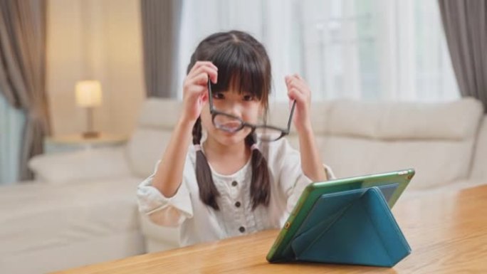 亚洲迷人的小可爱孩子坐在桌子上，看卡通或在家里的客厅平板电脑上学习。年轻的女学生戴眼镜保护视力从数字