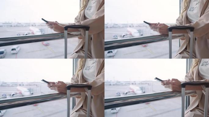特写女商人的手在等待航班时用手机发短信聊天，站在机场航站楼内商务旅行，手机和旅行行李以及飞机的背景
