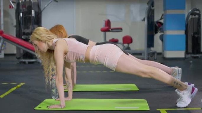 苗条身材的白人女运动员站在健身房的板形体垫上的侧视图。两个自信的年轻美女在室内锻炼。运动和锻炼概念